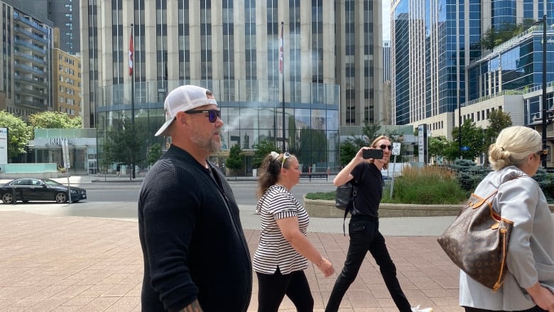 A man in a black sweater, white ballcap worn backwards, and sunglasses walks outside on a city street, surrounded by a few other people.