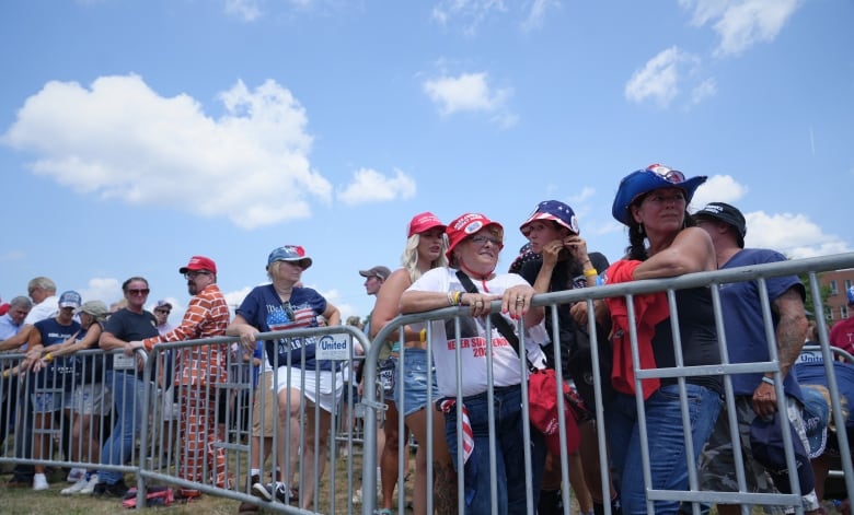 People wearing T-shirts supporting Donald Trump line up behind a barricade on a sunny day.