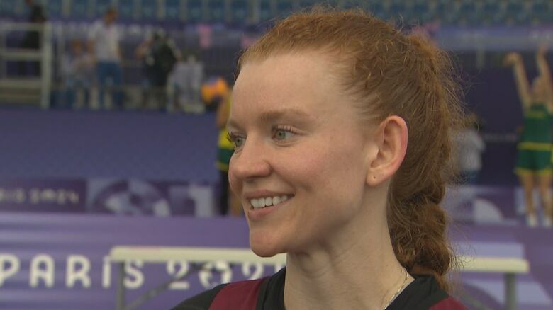 A white woman with red hair, tied in a braided pony tail, is smiling while wearing red basketball practice uniform. She is standing on a basketball court.