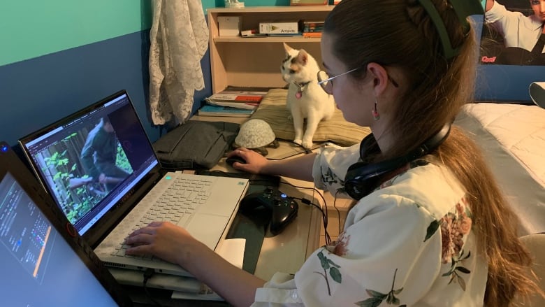 A young woman with a half-up half-down hairdo, wearing glasses, editing video on her laptop in her bedroom.