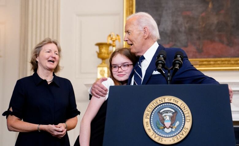 President Joe Biden, standing behind a podium, sings as he puts his arm around a teenage girl, with long hair and glasses. A woman stands behind,singing.