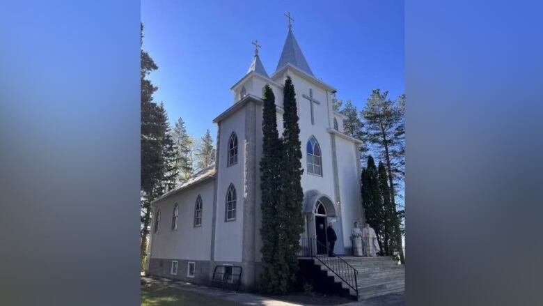 A white church with silver colour roof stands under a blue sky and beside tall trees.