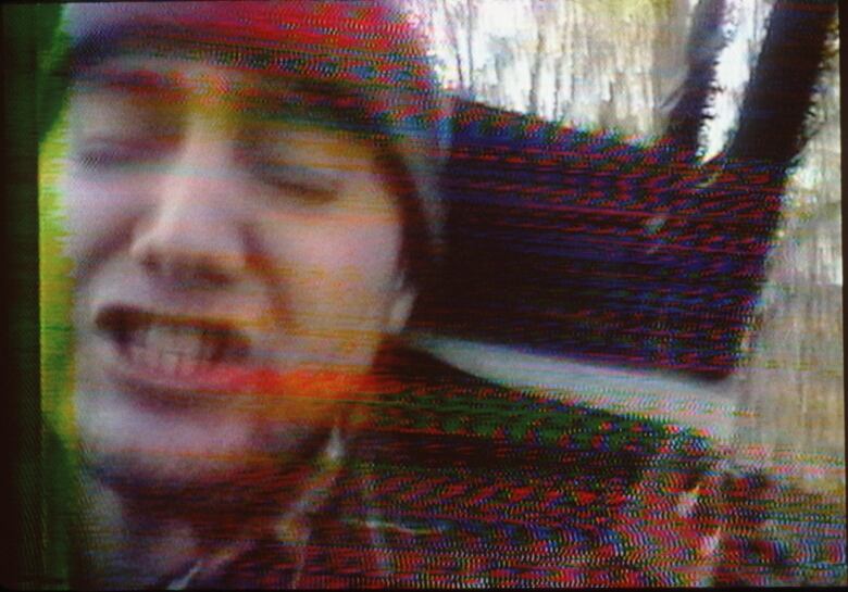 A man grimaces while standing outside in the woods. The image has blurry scratch marks as if seen on an old, damaged VHS tape.