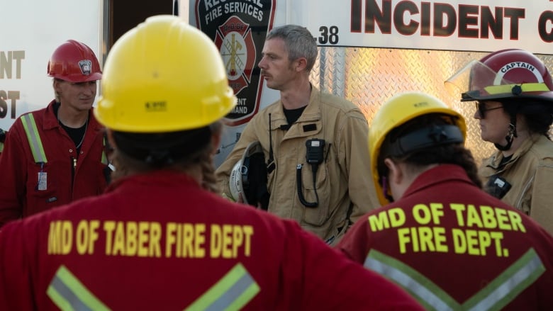 A group of men and women in helmets and coveralls gather around a speaker. 