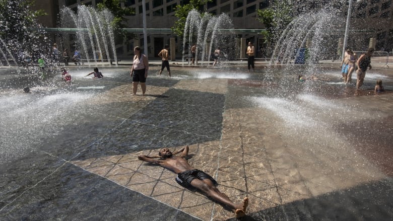 A man wearing shorts lies in a shallow spray pool outside with water splashing around him and people in bathing suits in the background.