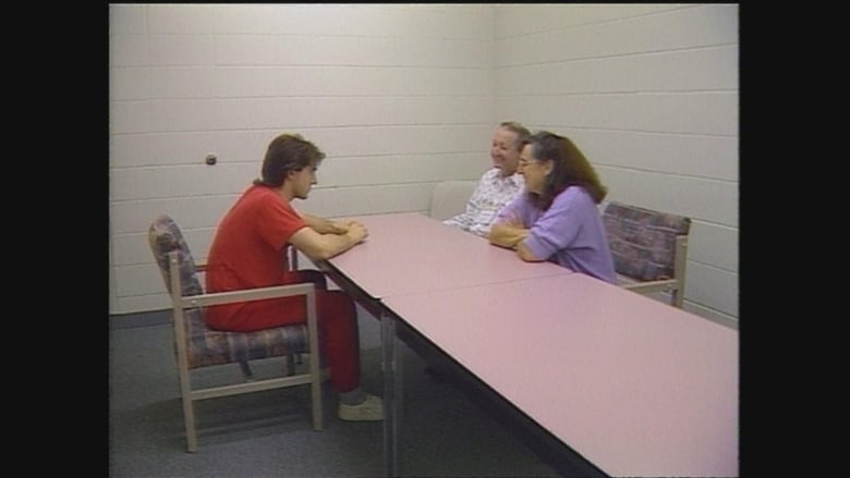 A young man in an orange jumpsuit sites at a table with a middle-aged woman and man sitting across from him. 