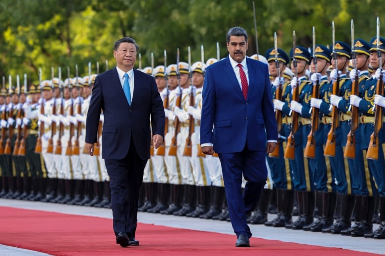 Tow men in suits walk down a red carpet as a line of soldiers holding rifles stand on guard.