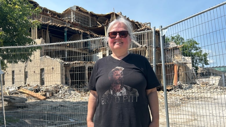 A woman with grey hair, wearing a black shirt and sunglasses, is standing in front of a fence surrounding a partially demolished building.