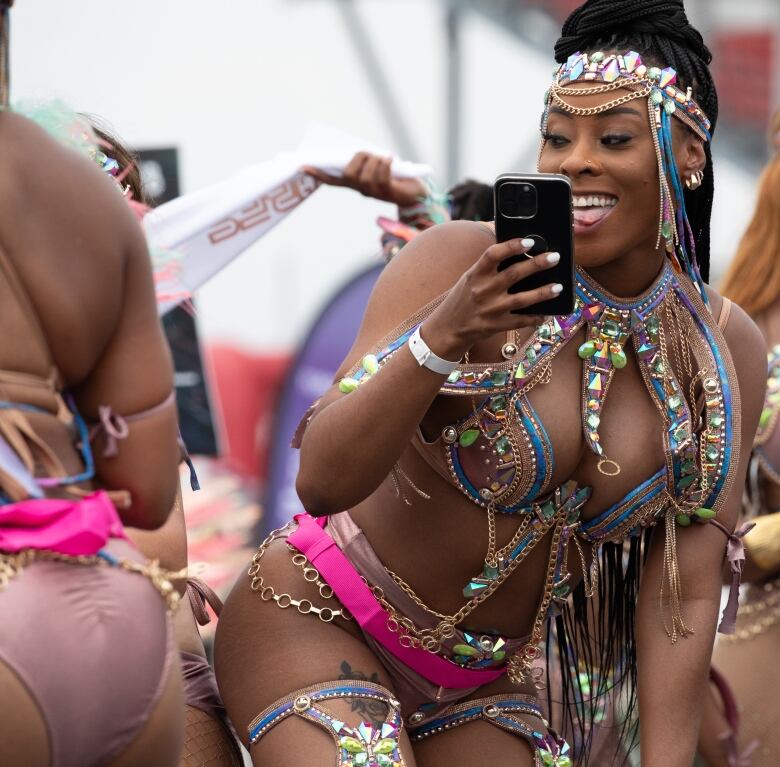 A woman in a festive, bedazzled bikini dances next to another woman just out of frame. She is holding her phone to take a video. The two are in the Carnival parade