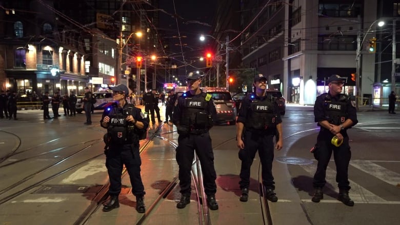 Photograph showing four police officers in a downtown toronto intersection, with more officers and police cars behind them. 