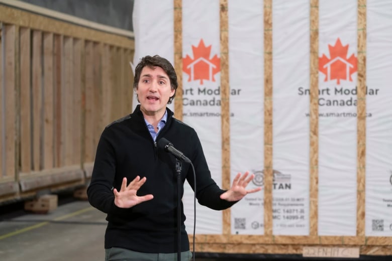 A white man gestures as he speaks in front of a row of prefabricated houses.