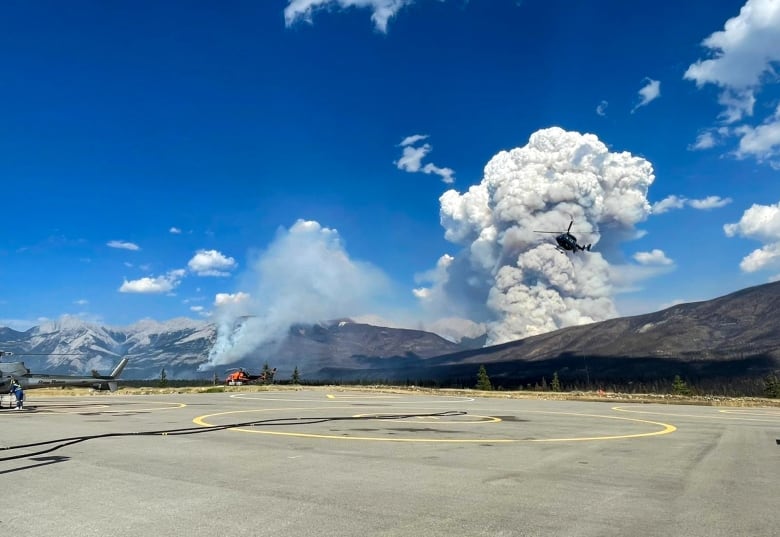 A helicopter makes its way to a landing pad as smoke billows in the background.