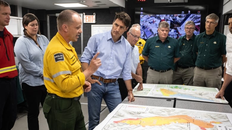 Wildfire Incident Commander Landon Shepherd, third from left and in a Parks Canada uniform, speaks with Prime Minister Justin Trudeau, who is wearing a blue shirt and blue jeans. They are in a small room with one black wall, which is the wildfire command centre in Hinton, Alberta.