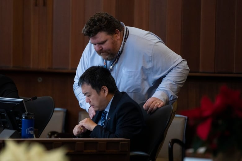 A man looks over at another man's screen in city council chambers.