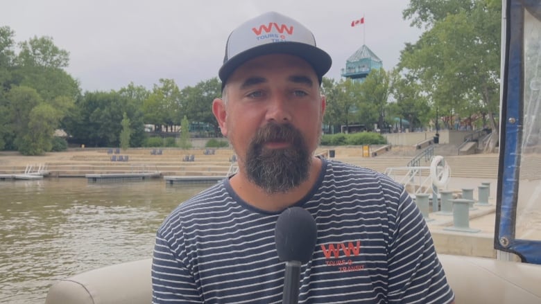 A man in a baseball cap sits in a boat and talks to someone.