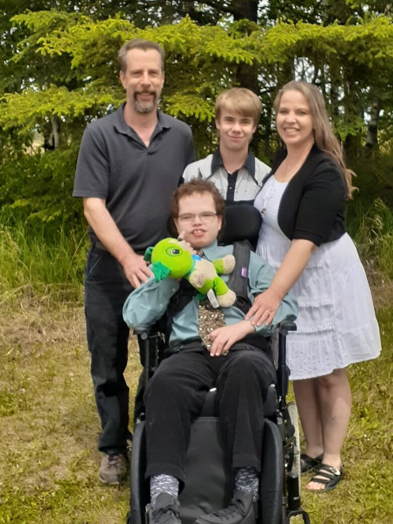 A family of four smiles outside with one son in a wheelchair holding a stuffed animal.