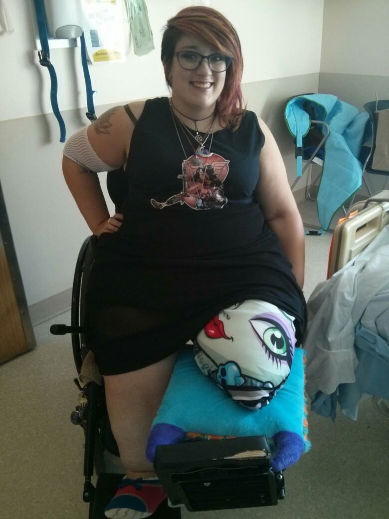 A person smiles with one hand on their hip as they sit in a wheelchair and has their left leg amputated. 