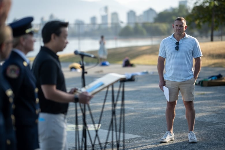 A person speaks at a podium outside as a man in shorts watches from the side. 