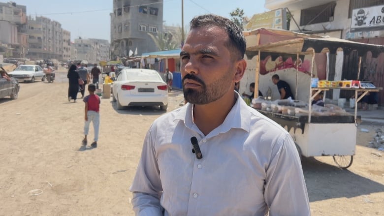 A man stands in front of a market wearing a grey collared shirt.