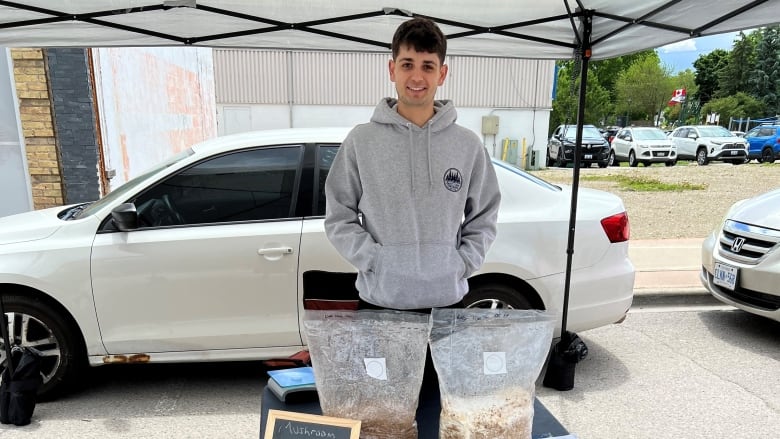 A young man in a grey hoodie standing in front of a black table with mushrooms in bags on it and a small chalkboard sign advertising the product for sale