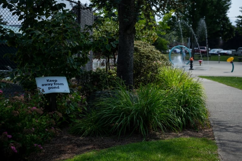 A sign reading 'keep away from pond' is seen surrounded by foliage and beside a fence with a waterpark nearby.