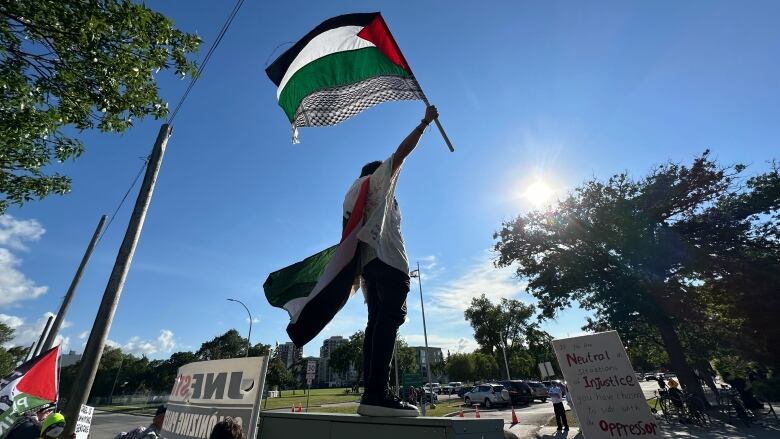 A person stands on an electrical unit box outside waving a Palestinian flag at a protest.