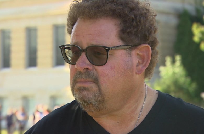 A man wears black sunglasses and stands outside while doing an interview with a reporter.