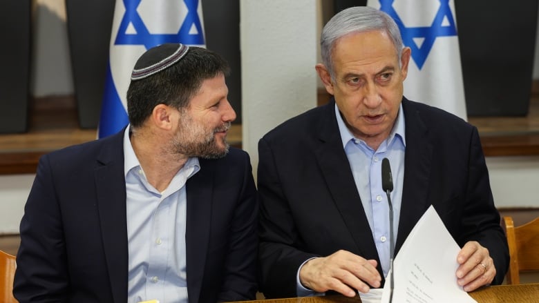 Two men sitting in front of Israeli flags talk at a table.