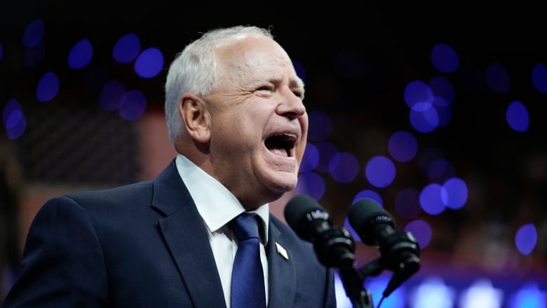 An older man with short white hear and wearing a suit has a wide open-mouth smile while standing at a podium