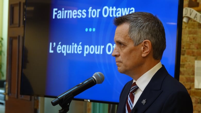 A mayor listens behind a microphone. A sign behind him says 'Fairness of Ottawa.'