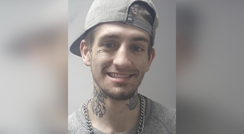 Smiling man with neck and face tatoos and a ballcap on backwards smiles at the camera.