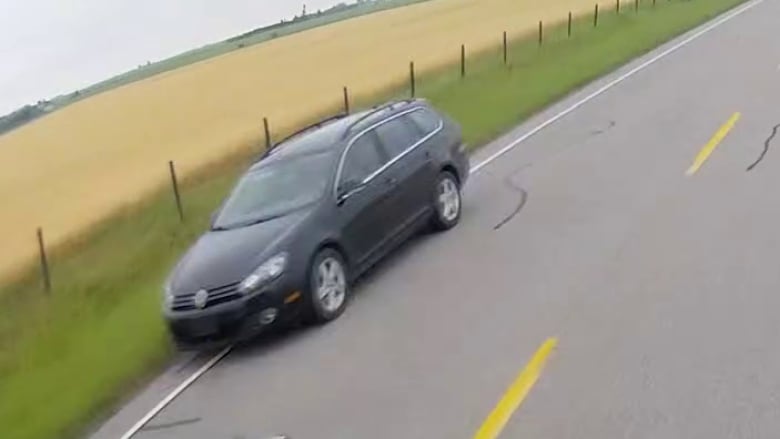 A dark vehicle driving down a rural road with a farmer's field on one side. 