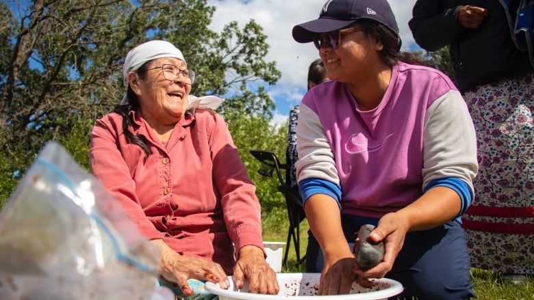 Two women kneel on the ground crushing choke cherries.