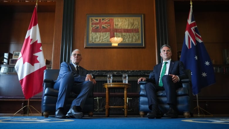 Two men in suits sit in arm chairs, flanked by the Canadian and Australian flags. 