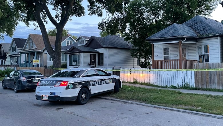 A police car is parked partly on a curb and boulevard in front of a house. Yellow police tape surrounds the fence in front of the house.