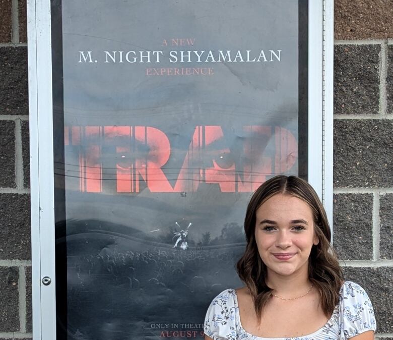 Juliet stands in front of a brick wall, with a movie poster for the movie Trap, directed by M Night Shyamalan, which she appears in.