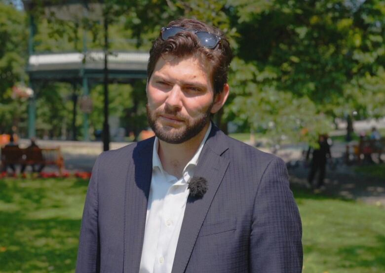 Man, wearing blue blazer and dress shirt, standing in park and looking at camera with neutral expression.