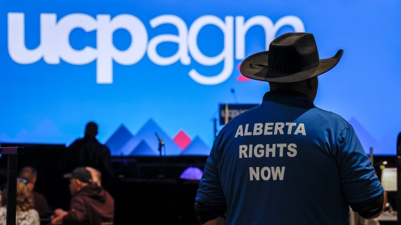 A man in a black hat with his back to the camera, lettering on his shirt.