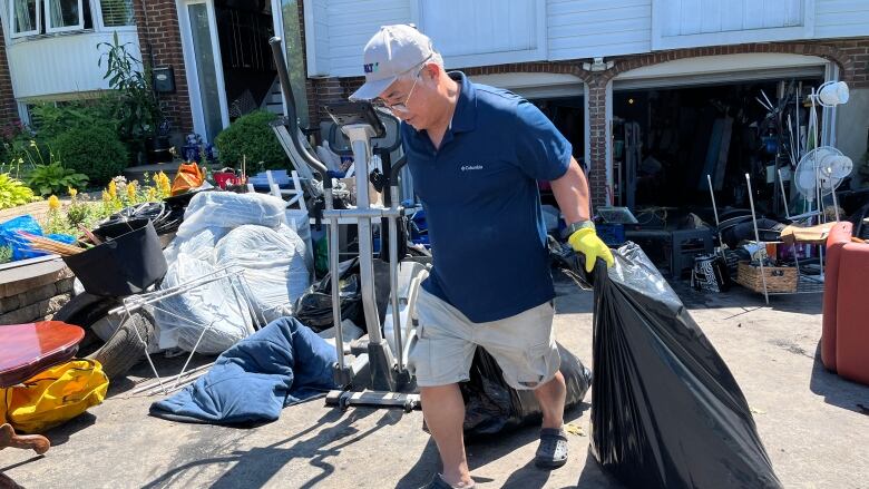 A man carries a bag of garbage to the curb as he cleans a basement post-flood. There are multiple items strewn across his garage driveway. 