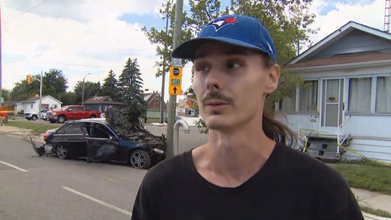 A young man with a mustache and blue ball cap is seen from the shoulders up in front of a wrecked car that's crashed into a pole on a city street next to a house in broad daylight in summer