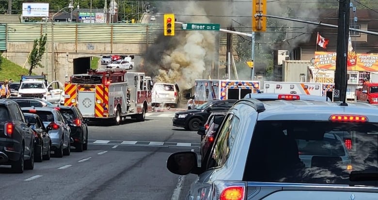 At a city intersection, fire crews work on a mini van with smoke coming from it. Traffic is backed up behind the scene. A chip truck is visible parked right next to the intersection on the far right corner. It's day time in the summer