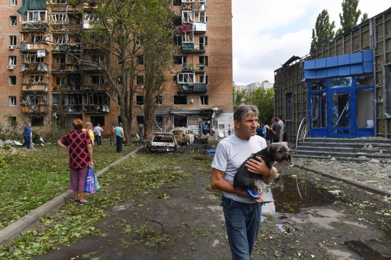 A man holding a dog walks near a damaged apartment building.