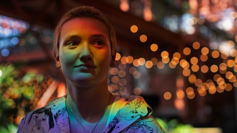 The face of a woman, with short blonde hair, is lit up in rainbow colours. She is wearing a graphic zip-up hoodie.