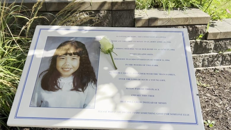 A plaque with an East Asian girl's picture on it in a park, with text honouring Mindy Tran and asking community members to remember her.