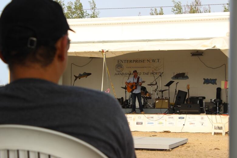 In the blurry foreground, the back of a man's head. In the focused background, a man playing guitar on a stage. 