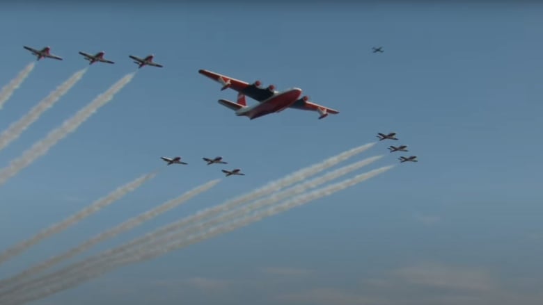 A massive water bomber escorted by nine fighter jets ejecting plumes of smoke into the sky.