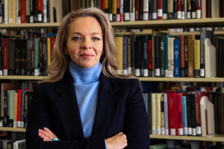 Woman with blond hair and blue shirt and black jacket with arms folded. Books are behind her.