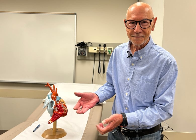 A 77-year-old man holding a piece of a pacemaker, standing beside a model of a heart