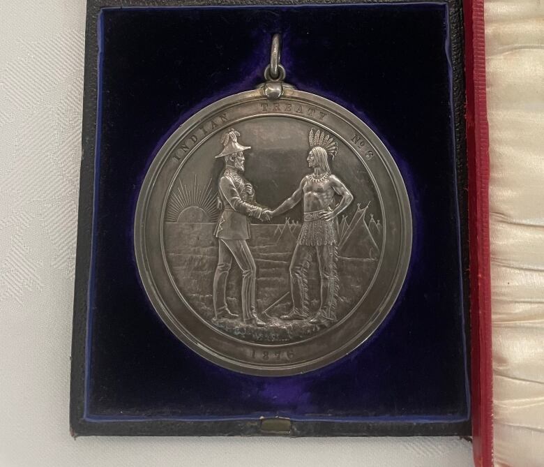 The silver medal sits in a blue velvet case. The medal shows a man in a headdress shaking hands with a British official against a background with a sun and teepee. 