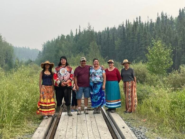 Six people stand side by side on railway tracks in a forest.